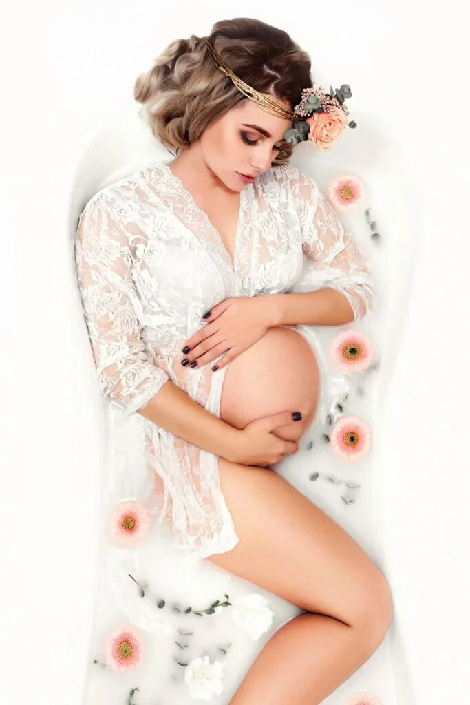 A pregnant woman cradles her belly in a milk bath with pink flowers.