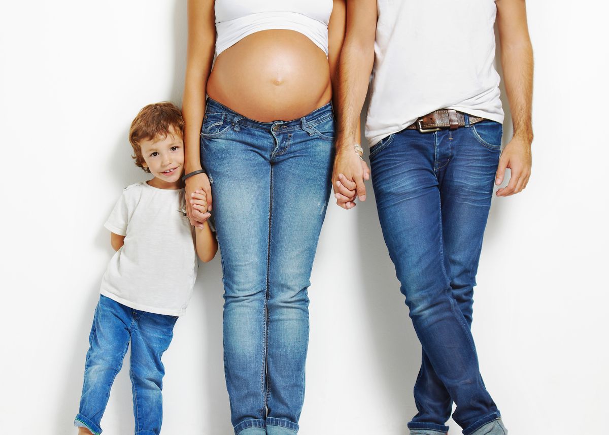 A family including a pregnant woman, a man, and a young boy pose in front of a white background while wearing jeans.