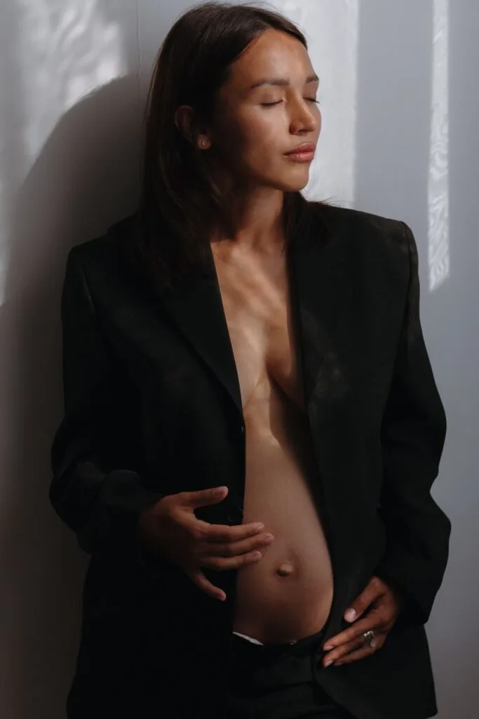 A pregnant woman in a black blazer with an exposed belly poses in low light.