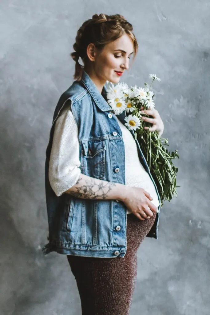 A pregnant woman in a jean jacket holds flowers over her belly.
