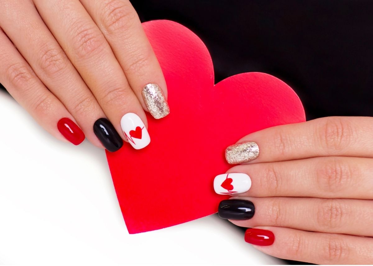 Hands hold a red cut-out heart with painted red, black, white, and silver fingernails with red hearts on the fingers painted white.