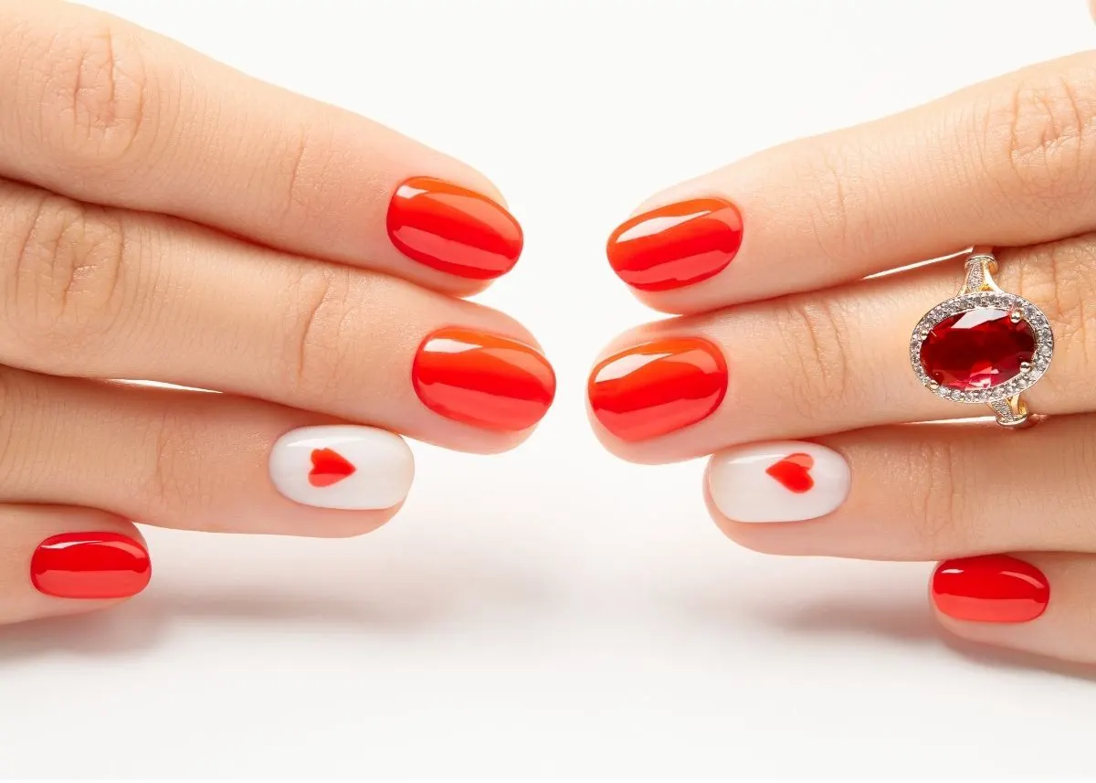 A woman shows off painted red fingernails with one white fingernail on each hand and a heart design on a white background.