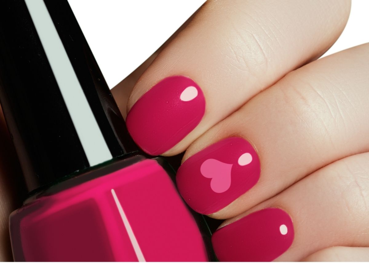A hand holds a bottle of pink fingernail polish with fingernails painted dark pink and one finger having a lighter pink heart design.