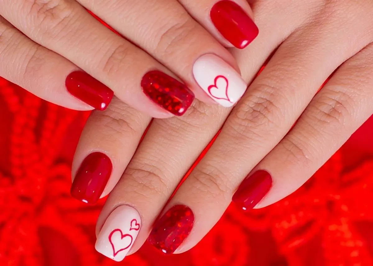 A hand rests on top of another hand, with fingernails painted red and one finger on each hand painted white with a red heart design.