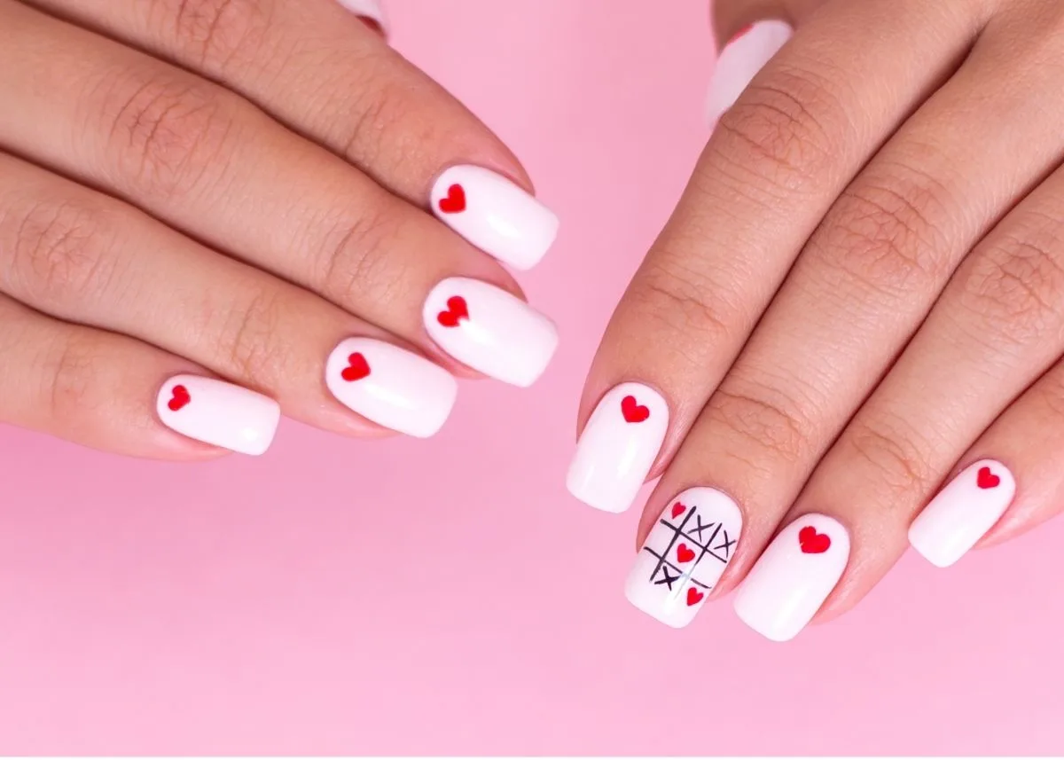 Hands rest on a pink background with painted white nails and a mini red heart above the cuticle of each finger.