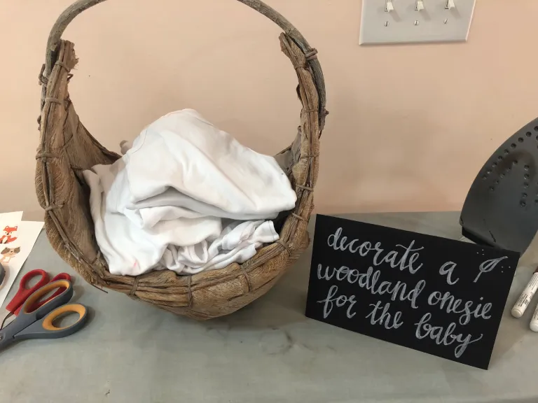 White onesies in a basket with a sign instructing baby shower party guests to decorate a onesie.