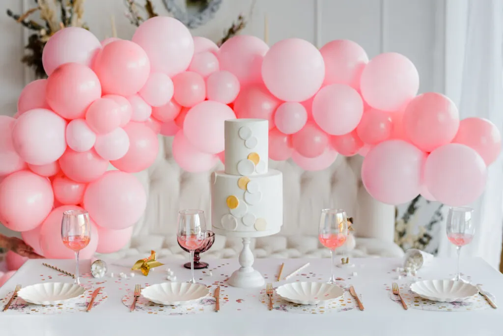 Table decorated with place settings and two-tier cake. Pink and white balloon garland in background.