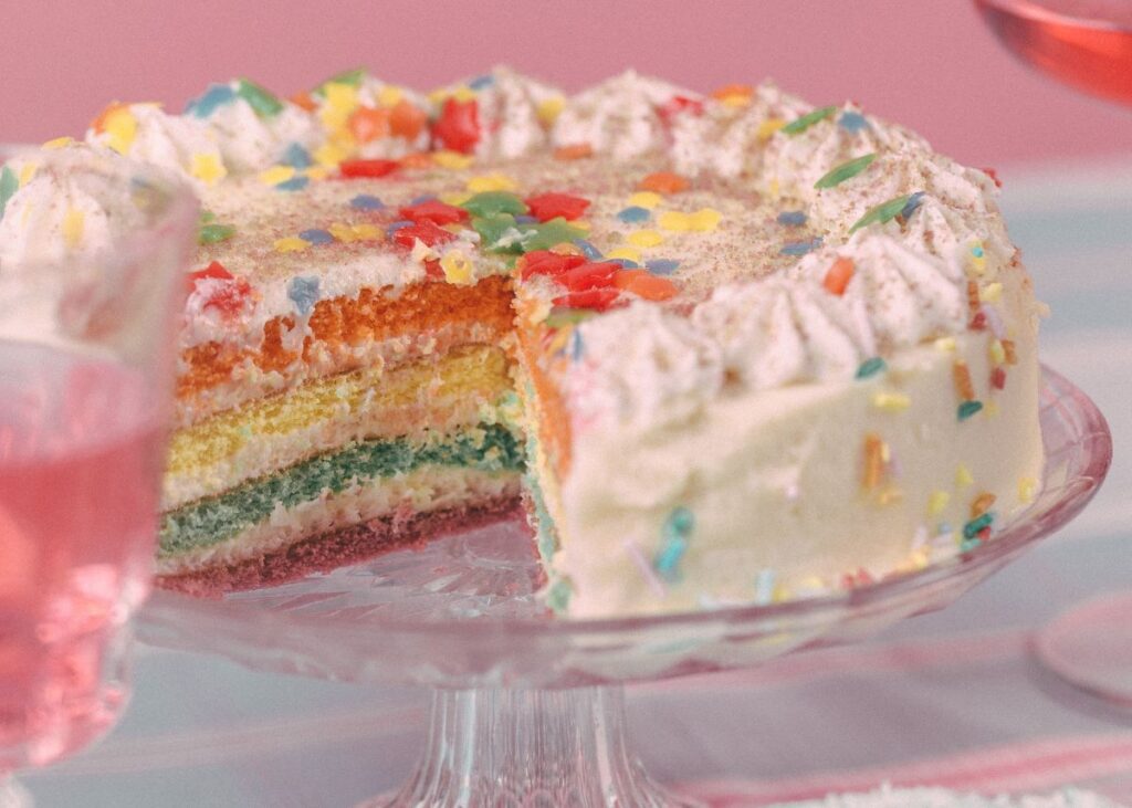 A rainbow cake with white icing and sprinkles with a big slice cut out.