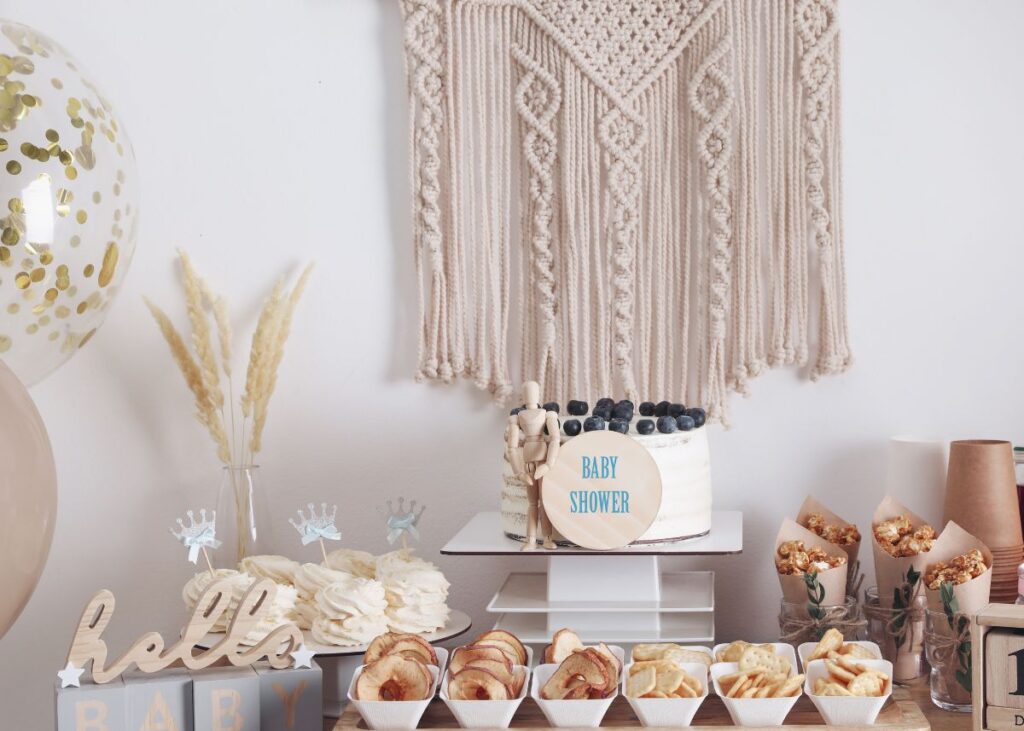 A dessert table at a baby shower with boho decor including a wall hanging and ornamental grass.
