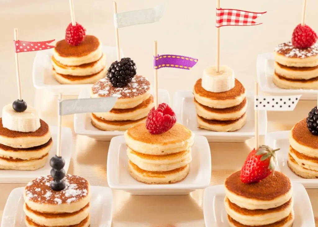 Mini pancakes stacked on white plates with cocktail flags.
