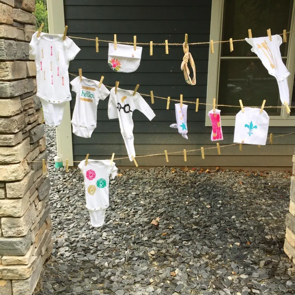 White baby onesies with painted on decorations hang from a clothesline outside.