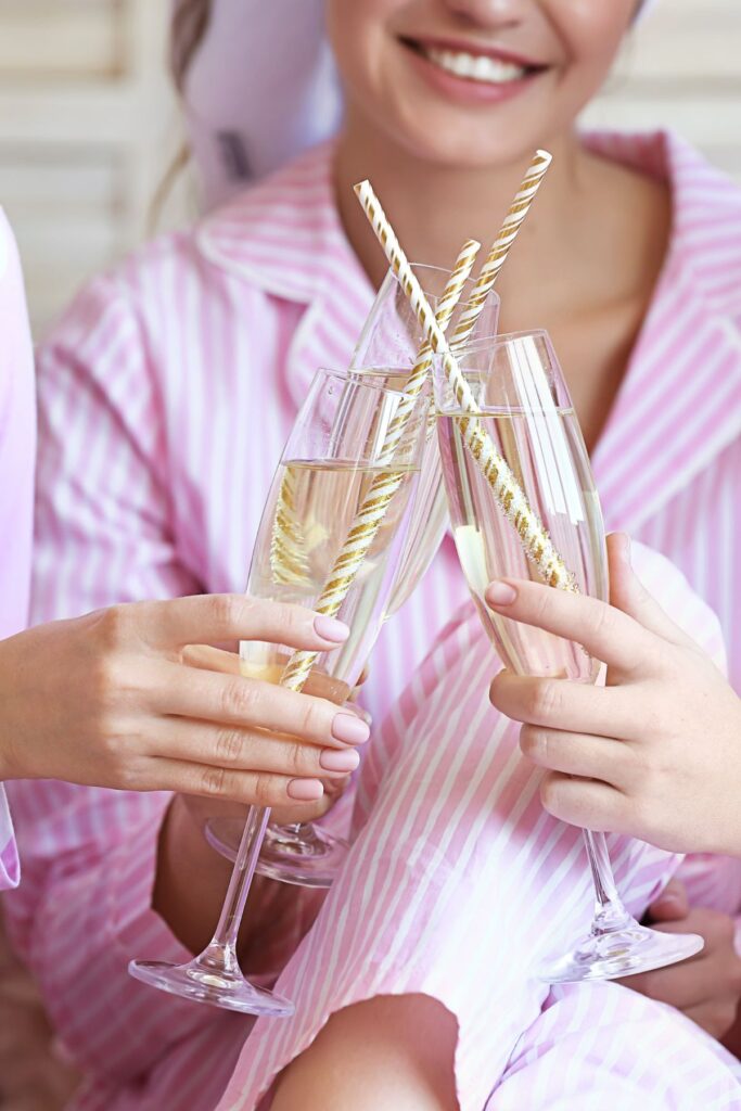 Women in pink pajamas clink champagne glasses with gold and white striped straws.