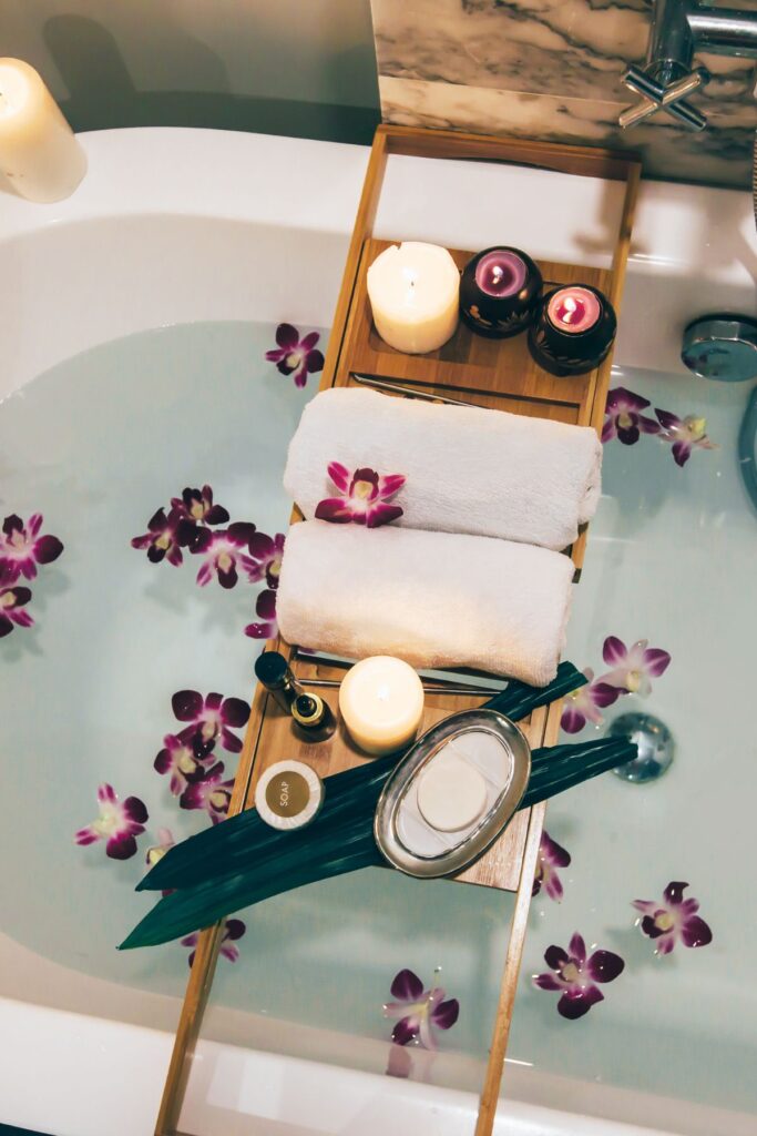 A bathtub filled with water and flower petals with a tray sitting over the top that has candles and towels on it.