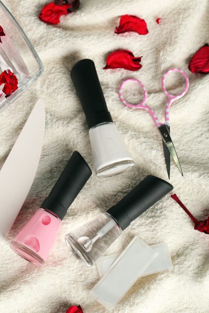 Pink, clear, and white nail polish on a white cloth with manicure tools and rose petals.