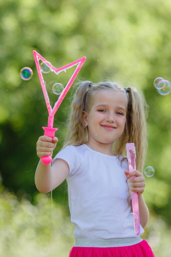A girl plays with a pink bubble wand outside with bubble floating in the air.