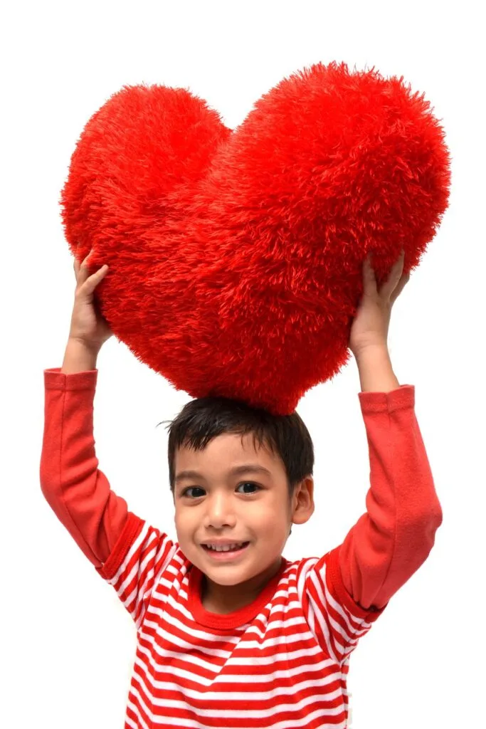 A boy in a red and white striped shirt holds a large red heart pillow over his head.