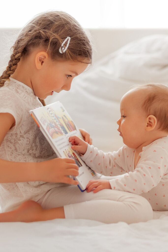 A young girl reads a book to her baby sister on a bed.