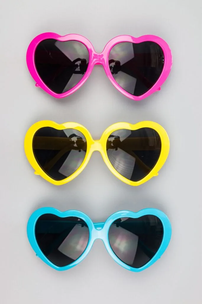 Three pairs of hear-shaped sunglasses in pink, yellow, and blue.
