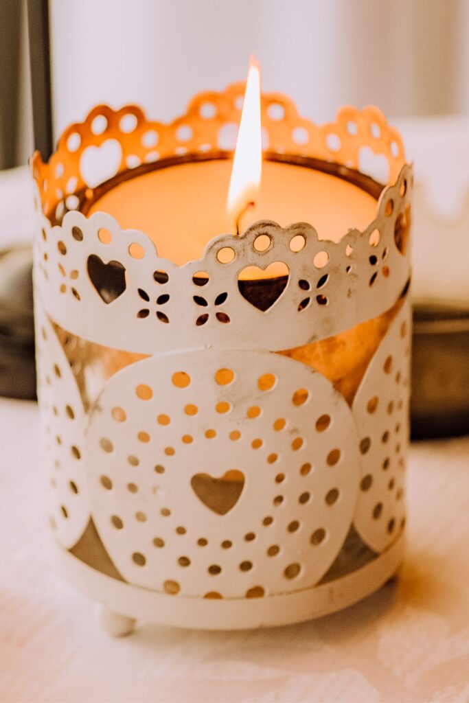 A candle burns inside a white metal candle holder with a lacey heart design.