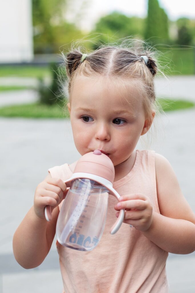 A toddler girl drinks from a clear sippy cup with a pale pink top.
