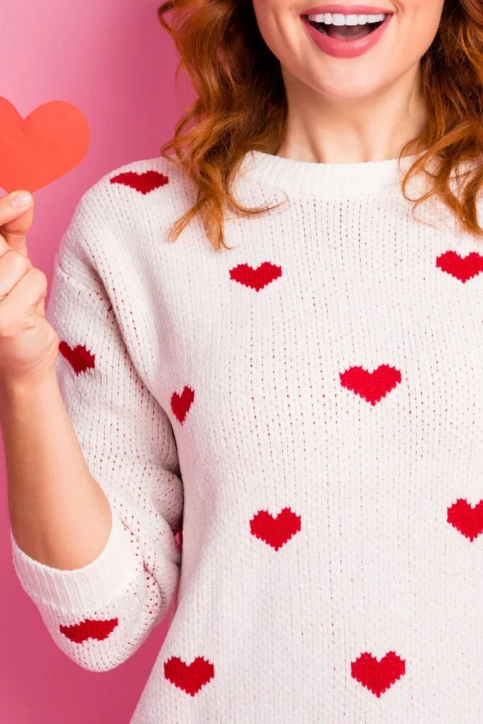 A woman in a white sweater with red hearts holds a red heart cut-out in her hand.