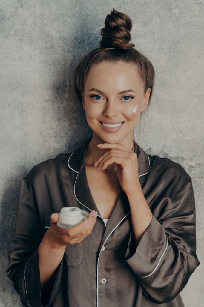 A woman in a darky gray silk robe holds a container of face cream.