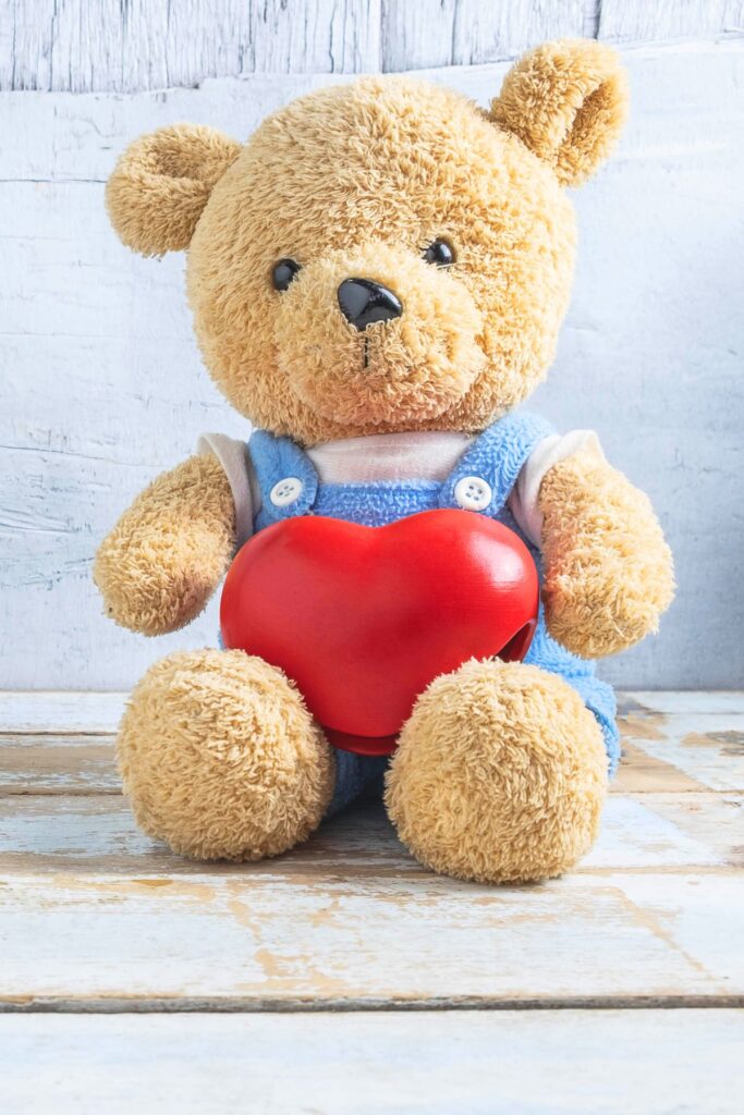 A teddy bear with blue overalls with a heart in its lap.