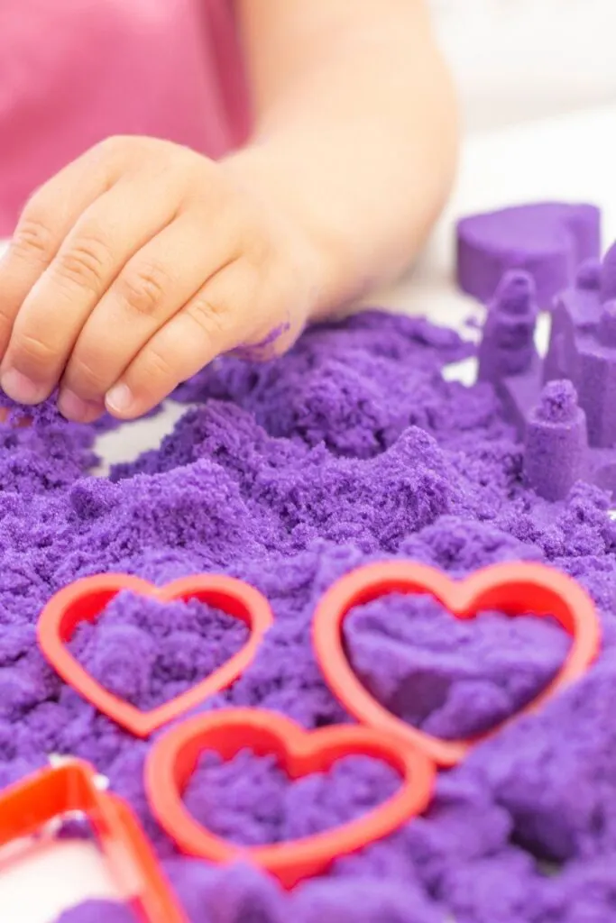A small hand plays with purple kinetic sand and heart-shaped molds.