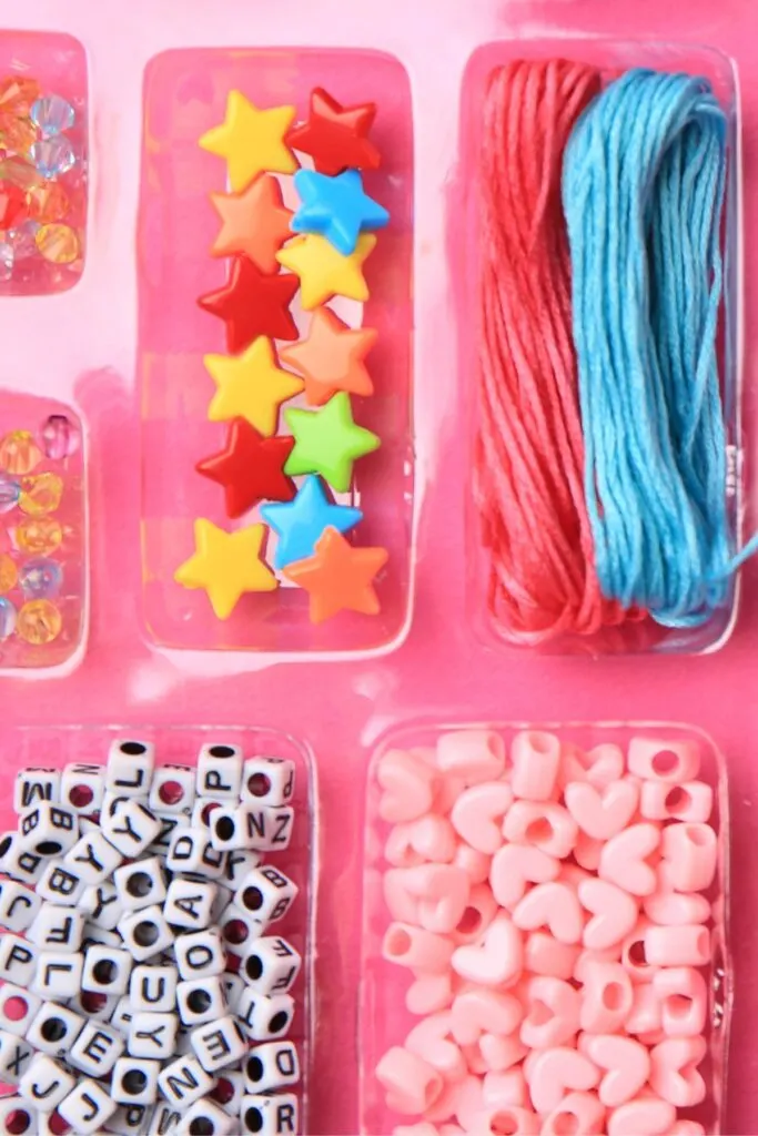 A jewelry-making kit for kids with various beads and strings on a pink background.