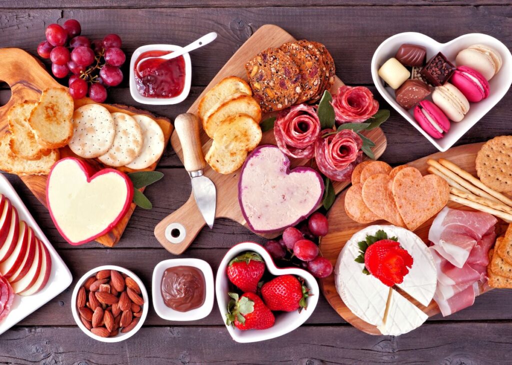 Grazing board with heart-shaped containers, charcuterie, cheese, and crackers.