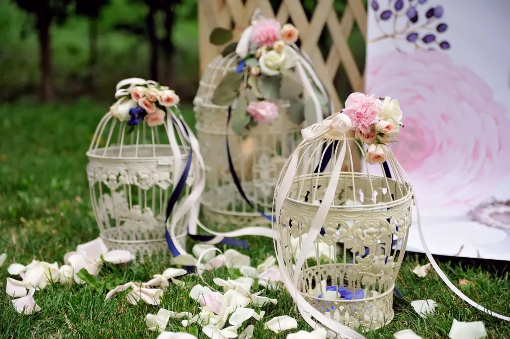 Bird cages sit on the ground outside that are decorated with ribbons and flowers.