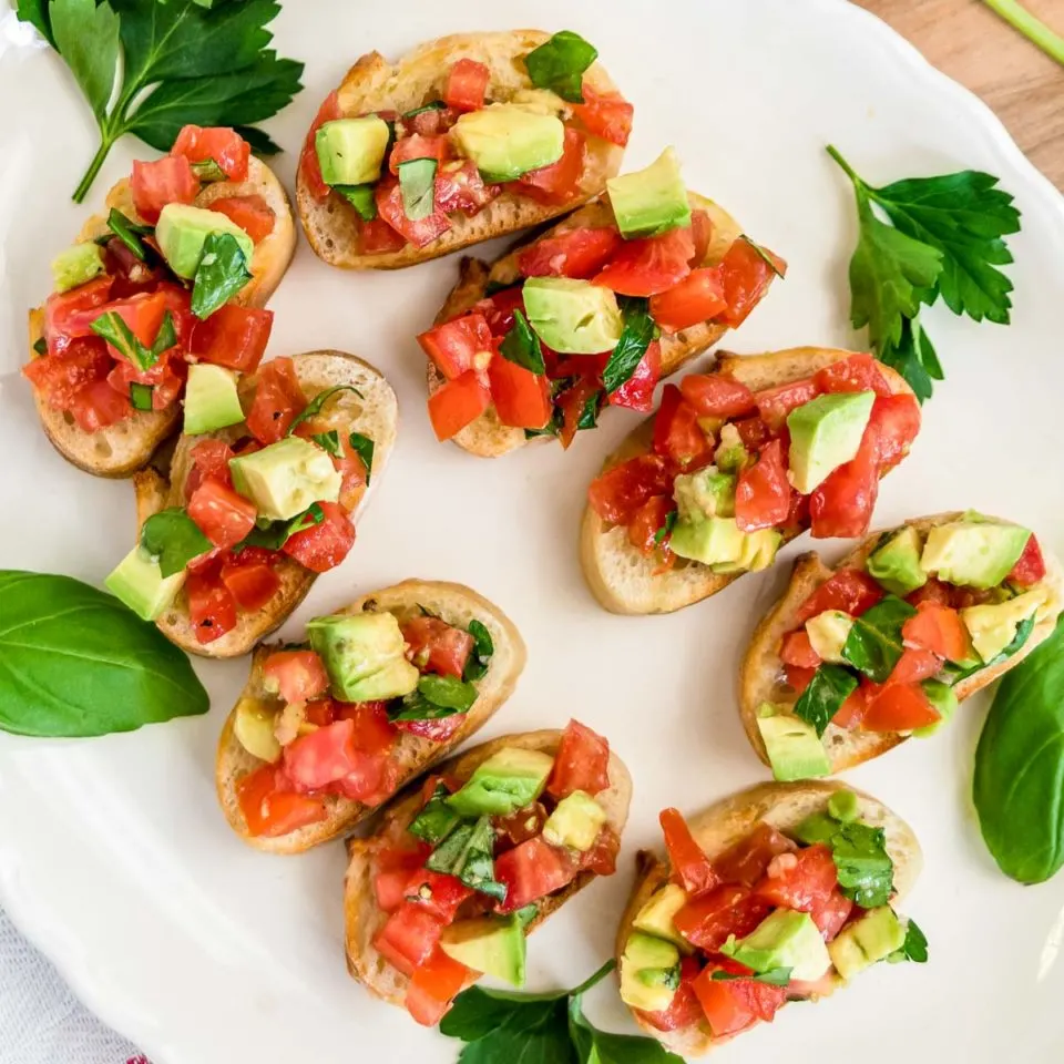 Tomato bruschetta with avocado on a white serving platter with green garnish.