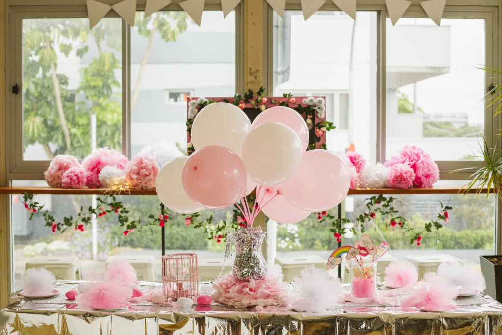 Balloons and flowers decorate a baby shower food table in front of a large window.