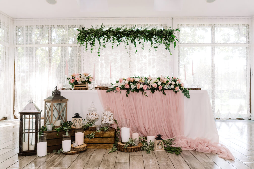 Flowers, lanterns, and greenery decorate a table with pink and white drapey tablecloths.