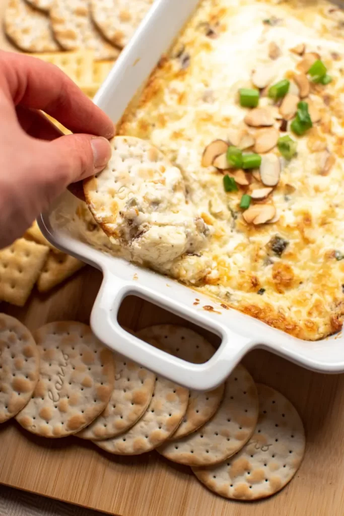 Toasted almond and Swiss cheese dip with a woman taking a scoop with a cracker.